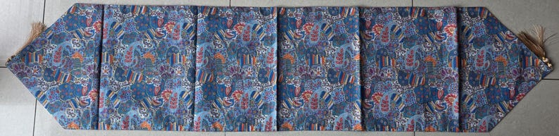 May include: A long, rectangular table runner with a blue, red, and white paisley pattern. The runner has a pointed end on each side with a tassel. The runner is made of fabric and is designed to be placed on a table.