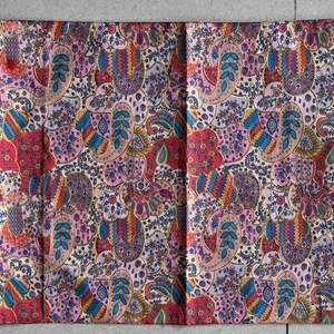 May include: A decorative table runner featuring a paisley design in red, purple, blue, and beige. The runner is rectangular with angled ends and gold tassels. The fabric appears to be quilted, with a length of approximately 180 cm.