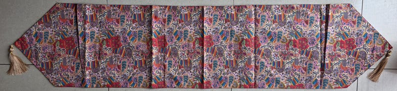 May include: A decorative table runner featuring a paisley design in red, purple, blue, and beige. The runner is rectangular with angled ends and gold tassels. The fabric appears to be quilted, with a length of approximately 180 cm.