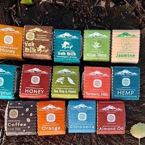 Puede incluir: Un surtido de jabones rectangulares con varias etiquetas, incluyendo "Sandalwood Honey", "Yak Milk", "Aloe Vera" y "Jasmine". Los jabones están dispuestos sobre una superficie de madera rústica, con una estética natural.