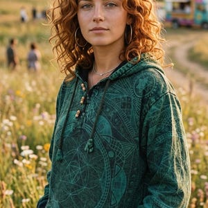 Könnte beinhalten: Eine Person trägt einen dunkelgrünen Kapuzenpullover mit geometrischem Muster in einem Feld mit Wildblumen. Der Kapuzenpullover hat eine Kapuze mit Kordelzug und Holzknebel. Die Person hat rote Locken und silberne Creolen.