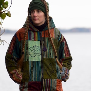 Peut inclure: Veste patchwork multicolore à capuche et fermeture éclair. Elle présente divers motifs et un patch vert avec le symbole Om. La personne porte un bandeau vert.