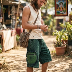 May include: A man wearing a cream-colored tank top, green shorts with a circular design on the pocket, and a brown leather shoulder bag. He is also wearing bracelets and a necklace. The scene is outdoors, with a market in the background.