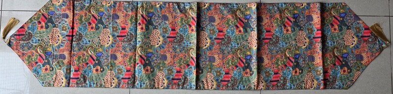 May include: A vibrant batik table runner. The fabric displays a mix of red, blue, green, and orange patterns. The runner is rectangular with angled ends and gold tassels. It would measure approximately 180 cm in length.