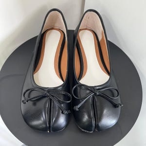 May include: A pair of black leather ballet flats with a bow detail on the toe. The shoes have a rounded toe and a low heel. The interior is lined with a light beige material.