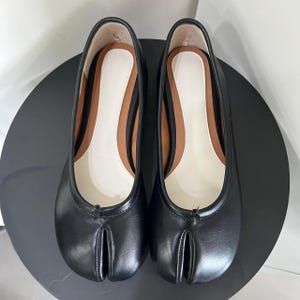 Op de afbeelding: Een paar zwarte leren ballerina's met een split-teenontwerp. De schoenen hebben een witte binnenzool met een bruine rand en een lage hak. De schoenen worden op een zwart oppervlak weergegeven.