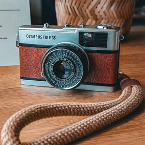 Op de afbeelding: Olympus Trip 35 camera met een bruine lederlook behuizing en zilveren bovenkant. De cameralens is zwart met een gedetailleerde diafragmaring. Een geweven bruine camerariem is voor de camera gedrapeerd.