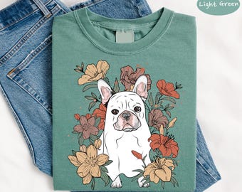 May include: A light green t-shirt featuring a white French Bulldog illustration surrounded by colorful flowers. The dog has a sweet expression. The shirt is folded on top of blue denim jeans.