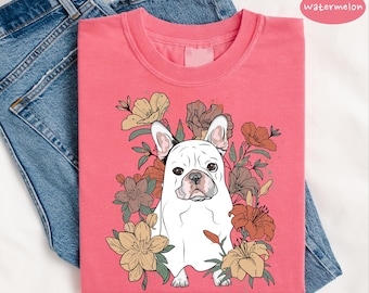 May include: A coral-colored t-shirt featuring a white French bulldog illustration surrounded by floral elements in shades of brown, yellow, and orange. The word "watermelon" is printed on the top right. The shirt is folded on a pair of blue jeans.