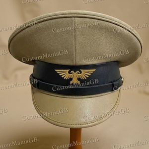 May include: A khaki-coloured military hat with a black band and a gold emblem. The hat has a rounded top and a visor. The hat is displayed on a wooden stand. The text "CustomManiaGB" is visible.