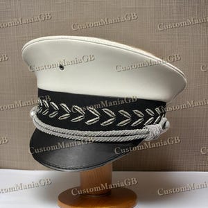 May include: A white and black military style hat with a silver braid and a black leather band. The hat has a white top and a black band with a silver braid around the brim.