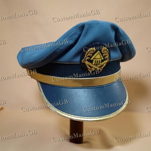 May include: A blue hat with a gold emblem and trim. The hat has a rounded top and a visor. The emblem features a gold design on a black background. The hat is displayed on a wooden stand.