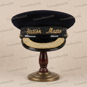 以下が含まれることがあります： 金色の刺繍とトリムが施されたネイビーブルーの「Station Master」ハット。木製のスタンドに展示されています。帽子は湾曲したバイザーと装飾的な金色のアクセントが特徴です。