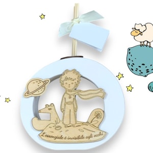 May include: A light blue circular wooden decoration featuring a Little Prince theme. The design includes a wooden cutout of the Little Prince, a fox, a planet, and the text "L'essenziale è invisibile agli occhi". A sheep is on a blue planet.