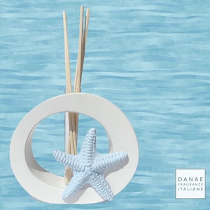 Peut inclure: Diffuseur en céramique blanche avec une ouverture ronde et une étoile de mer bleue. Le diffuseur contient des bâtonnets en bois et se trouve sur un fond rayé bleu et blanc. Le texte "DANAE FRAGRANZE ITALIANE" est dans le coin inférieur droit.