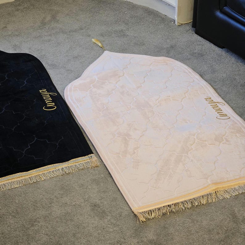 May include: Two prayer rugs, one black and one light pink, are displayed on a gray surface. The rugs have an arched top and a decorative pattern. The word "Camaya" is embroidered in gold on each rug, and they have a gold fringe.