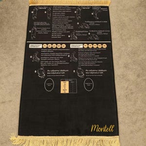 Op de afbeelding: Zwart gebedskleed met gouden franjes en witte instructiediagrammen. Het kleed toont Arabische tekst en illustraties van gebedsposities. De merknaam "Montell" is rechtsonder in goud gedrukt.