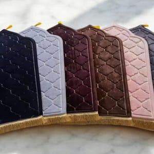May include: A collection of prayer rugs in various colors, including navy, gray, burgundy, brown, pink, and purple. Each rug has a decorative arched top and a geometric pattern. The rugs are displayed on a light-colored surface.