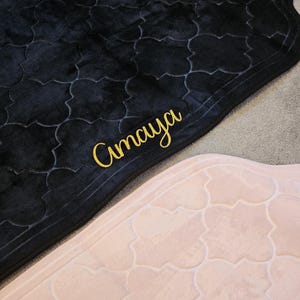 May include: Two prayer rugs, one black and one light pink, with a repeating quatrefoil pattern. The black rug has the word "Amaya" embroidered in gold script. The rugs are made of a soft, plush material.