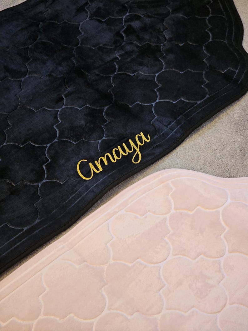 May include: Two prayer rugs, one black and one light pink, with a repeating quatrefoil pattern. The black rug has the word "Amaya" embroidered in gold script. The rugs are made of a soft, plush material.