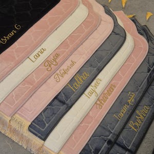 Op de afbeelding: Een collectie rechthoekige gebedskleden in zwart, crème, oudroze en donkergrijs, elk voorzien van een gestructureerd geometrisch patroon en goudkleurig geborduurde namen zoals Hassan G., Lana, Ayca, Nafesah, Talha, Tayseer, Shareen, Tanim en Bushra.