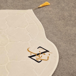 Puede incluir: Alfombra de oración color crema con un patrón geométrico y una borla dorada. La alfombra presenta caligrafía árabe negra y dorada. La alfombra está sobre una superficie gris.
