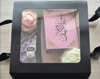May include: A black gift box with a clear window showing a pink and white rose, a small digital prayer counter, a string of beads, and a pink card with Arabic calligraphy that says "Happy Eid".