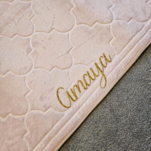 May include: A light pink, patterned rug with a repeating quatrefoil design. The word "Amaya" is written in gold glitter script in the lower right corner. The rug has a soft, plush texture and a light border.
