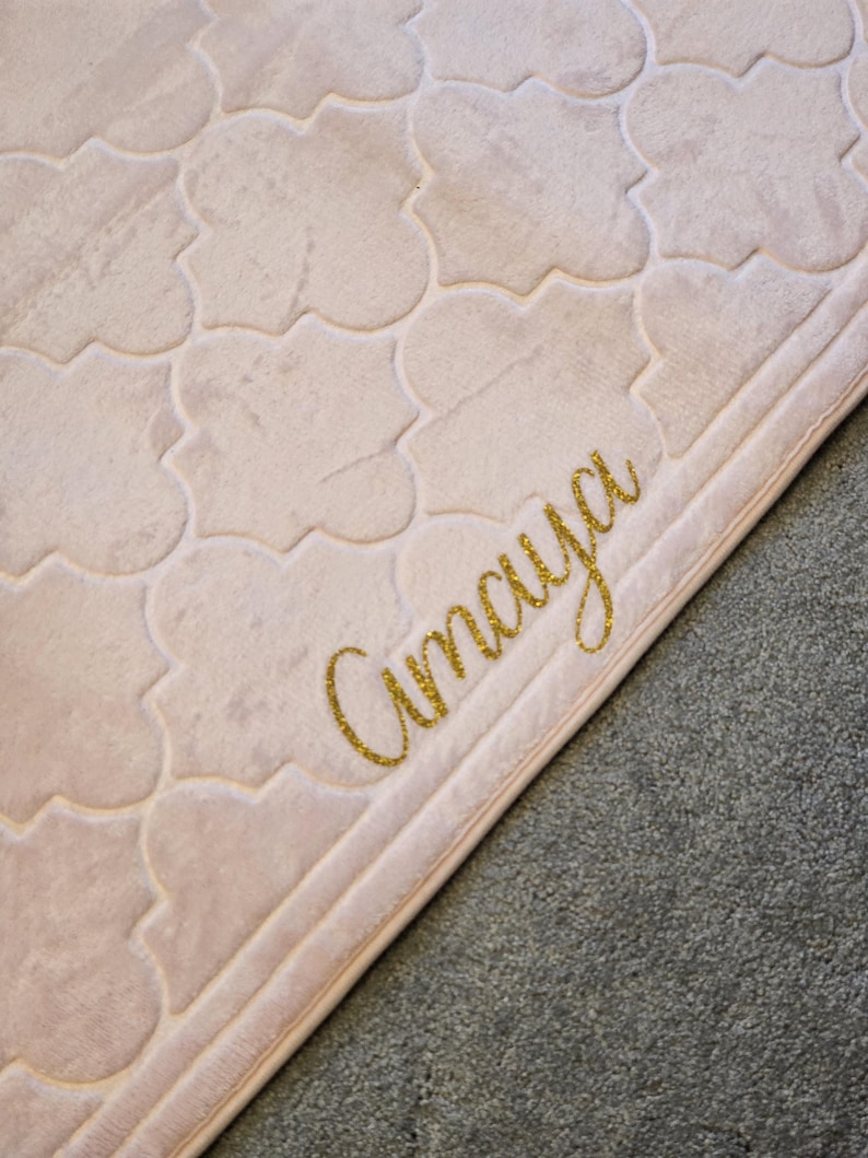 May include: A light pink, patterned rug with a repeating quatrefoil design. The word "Amaya" is written in gold glitter script in the lower right corner. The rug has a soft, plush texture and a light border.
