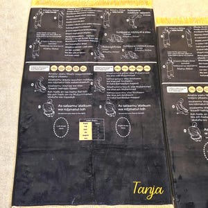 May include: Black prayer rug with detailed instructional diagrams and Arabic text. The rug features illustrations of prayer positions and phrases. The name "Tanja" is written in gold script at the bottom.