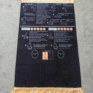 May include: Black prayer mat with gold fringe. The mat has white diagrams and text detailing prayer positions and phrases. The rectangular mat is designed for religious practice and has a decorative border.