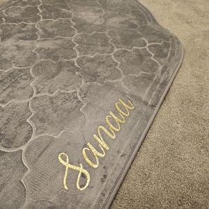 Può includere: Un tappeto da preghiera grigio con un motivo geometrico e il nome "Sanaa" ricamato in filo d'oro.