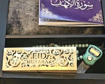May include: A black box with a gold-colored box that says "Eid Mubarak" inside. The box also contains a purple book with Arabic writing, a green prayer bead necklace, and a green digital prayer counter.