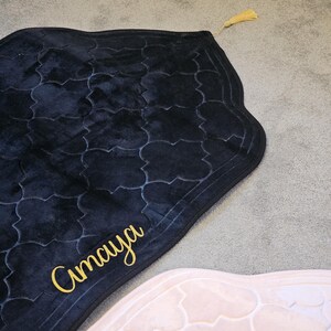 May include: A black prayer rug with a patterned design and gold tassel. The word "Amaya" is written in gold script on the rug. The rug is on a gray carpet.