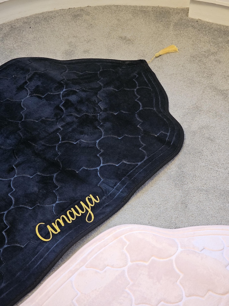 May include: A black prayer rug with a patterned design and gold tassel. The word "Amaya" is written in gold script on the rug. The rug is on a gray carpet.