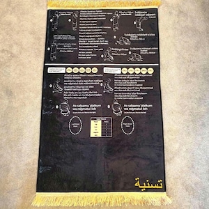 Puede incluir: Alfombra de oración negra con instrucciones y diagramas detallados en blanco. La alfombra tiene un fleco amarillo y texto en árabe. Los diagramas ilustran las posiciones de oración. La alfombra está diseñada para la práctica religiosa.