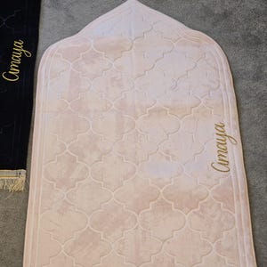 May include: A light pink prayer rug with a repeating quatrefoil pattern. The rug has a gold tassel at the top and gold fringe at the bottom. The name "Amaya" is written in gold script on the side.