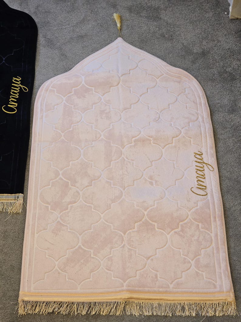 May include: A light pink prayer rug with a repeating quatrefoil pattern. The rug has a gold tassel at the top and gold fringe at the bottom. The name "Amaya" is written in gold script on the side.