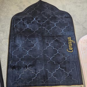 May include: A dark blue prayer rug with a geometric pattern and gold fringe. The rug has a decorative arch shape at the top and the name "Amaya" in gold script on the side. A gold tassel hangs from the top.