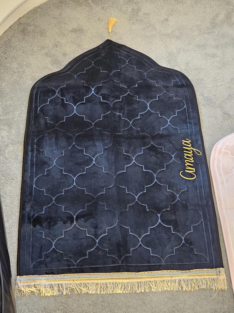 May include: A dark blue prayer rug with a geometric pattern and gold fringe. The rug has a decorative arch shape at the top and the name "Amaya" in gold script on the side. A gold tassel hangs from the top.