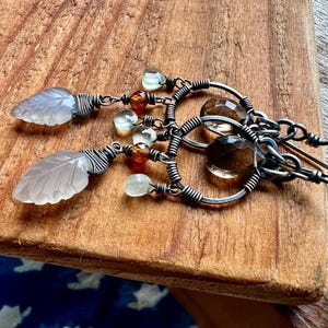 Op de afbeelding: Een paar zilveren oorbellen met draadgewikkelde bladeren, heldere en bruine edelstenen en een hoepelontwerp.