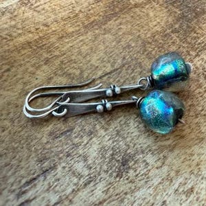 Puede incluir: Un par de pendientes colgantes de plata con cuentas de vidrio facetadas e iridiscentes. Las cuentas reflejan tonos de azul, verde y morado. Los pendientes se muestran sobre una superficie de madera.