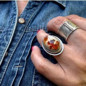 May include: A hand wearing two silver rings. One ring features a large, oval opal with orange and white patterns. The other is a wide band with a textured design. The rings are set against a blue denim jacket.