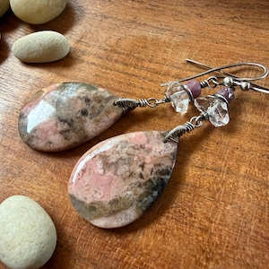 May include: A pair of drop earrings featuring large, polished, teardrop-shaped pink and gray mottled stones. Each stone is wrapped in silver-toned wire and connected to a small clear crystal bead and a tiny purple bead on a silver-toned hook.