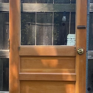 May include: A vintage wooden door with a glass panel and three solid wood panels below. The door has a brass doorknob and hinges. The wood has a warm, natural finish, and the door is set against a wooden fence.