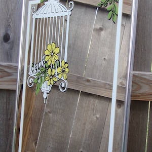 May include: A silver framed mirror with a white and green birdcage design with yellow flowers. The mirror is reflecting a wooden fence.