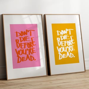 Op de afbeelding: Twee ingelijste prints met dezelfde tekst: "Don't die before you're dead." De print aan de linkerkant heeft een roze achtergrond en de print aan de rechterkant heeft een gele achtergrond.