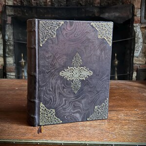K&ouml;nnte beinhalten: Ein dunkelbraunes, ledergebundenes Buch mit aufwendigen goldenen Metallverzierungen auf dem Einband. Das Buch hat ein marmoriertes Muster auf dem Einband und einen goldenen Quasten.