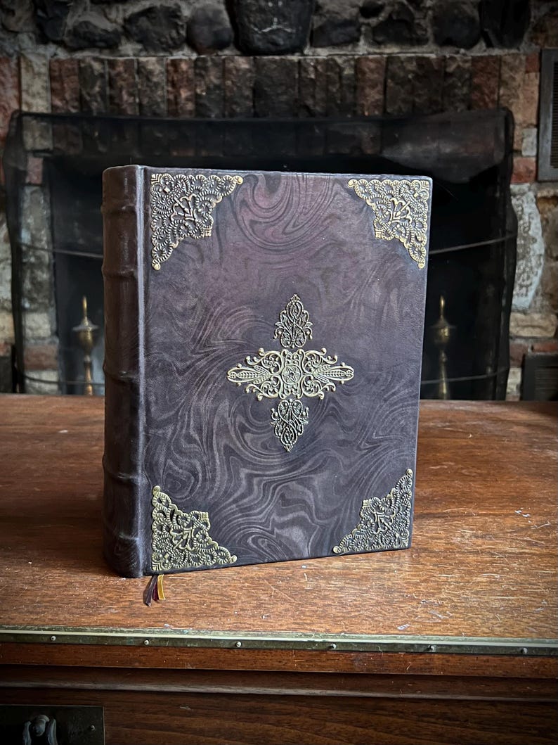 K&ouml;nnte beinhalten: Ein dunkelbraunes, ledergebundenes Buch mit aufwendigen goldenen Metallverzierungen auf dem Einband. Das Buch hat ein marmoriertes Muster auf dem Einband und einen goldenen Quasten.