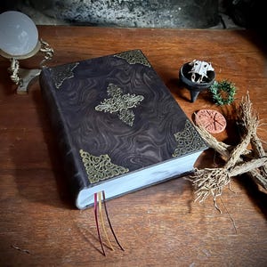 K&ouml;nnte beinhalten: Ein dunkelbraunes, ledergebundenes Buch mit goldenen Verzierungen und einem dekorativen Kreuzmuster auf dem Einband. Das Buch hat drei rote, goldene und wei&szlig;e Lesezeichen aus Band. Das Buch liegt auf einem Holztisch mit anderen Dekorationsgegenst&auml;nden.